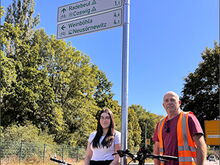 Auszubildende der Stadtverwaltung Coswig, Monique Liebetrau, und Steffen Wendler vom Baubetriebshof stehen an einem der neu angebrachten Radwegweiser an der Dresdner Straße.