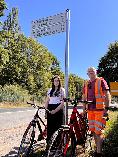 Auszubildende der Stadtverwaltung Coswig, Monique Liebetrau, und Steffen Wendler vom Baubetriebshof stehen an einem der neu angebrachten Radwegweiser an der Dresdner Straße.